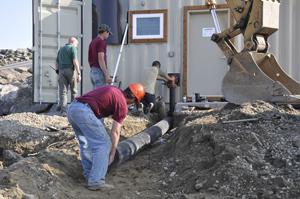 Workers at a remote site in Maine lay discharge pipe from a Lifewater Engineering system. The wood framing around the door enables the attachment of a 4- by 8-foot-long arctic entry. The framing around the windows enables the staff to screw on plywood sheets if flying ice or debris breaks the glass.